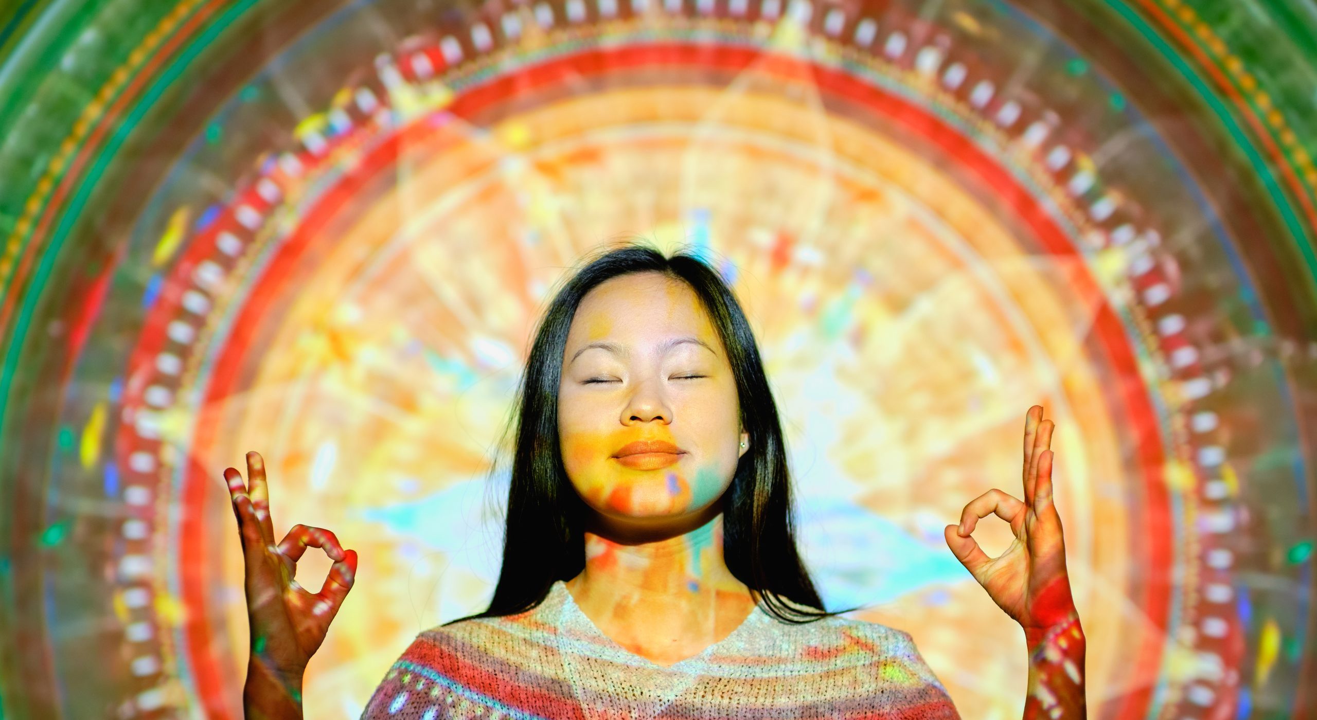 Content Asian female with closed eyes meditating with Gyan mudra hands near wall with bright glowing mandala in light studio